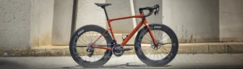 Una moderna bicicleta de carretera con cuadro rojo y ruedas de sección profunda, diseñada para el máximo rendimiento, se alza orgullosa contra una pared de hormigón.