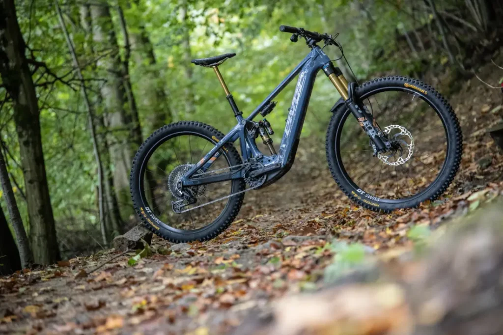 Una bicicleta de montaña Pivot Shuttle LT se encuentra en un sendero forestal cubierto de hojas, rodeado de árboles verdes.