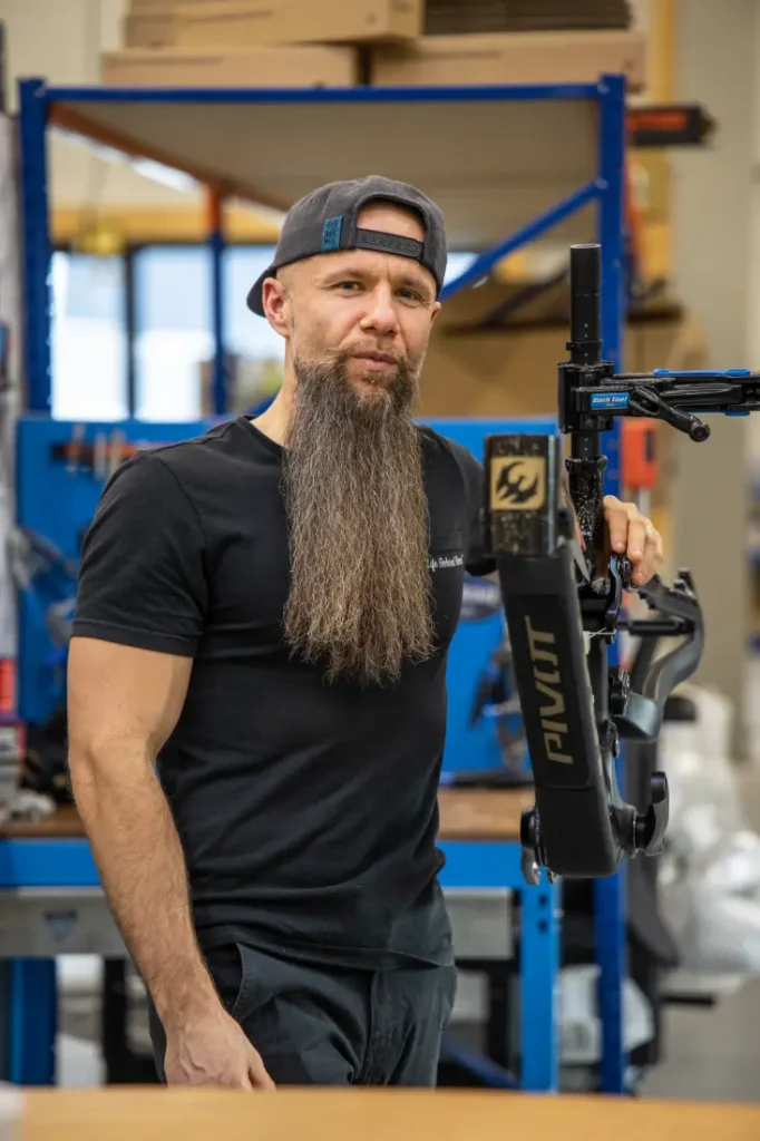 En un concurrido taller de Central Europea de Pivot, una persona con barba larga y gorra hacia atrás permanece de pie, con confianza, sosteniendo un cuadro de bicicleta.