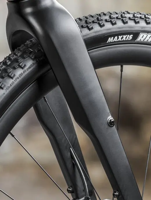 Primer plano de una horquilla y un neumático de bicicleta negros con bandas de rodadura texturizadas y la marca "Maxxis" en la pared lateral, combinados con elegantes horquillas de carbono para un mejor rendimiento.