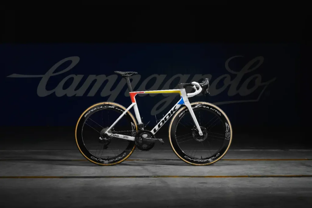 Una bicicleta de carretera blanca y azul con detalles en amarillo se destaca sobre un fondo oscuro con la palabra "Campagnolo" en la parte posterior.