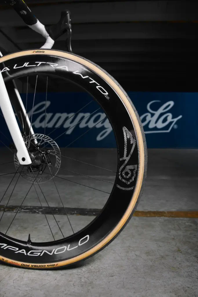 Primer plano de la rueda delantera de una bicicleta con llanta y neumático negros con el logotipo de Campagnolo. En el fondo se ve un cartel de Campagnolo borroso sobre una pared azul.
