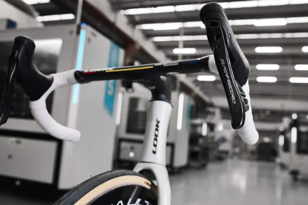 Primer plano de una bicicleta de carreras en un taller, con el manillar y el sillín en primer plano. Al fondo se ven máquinas y equipos industriales.