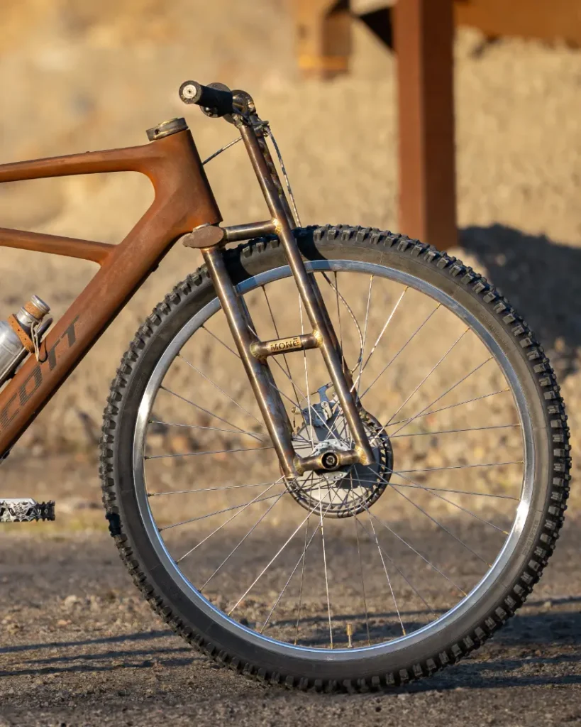 Primer plano de la rueda delantera de una bicicleta de montaña SCOTT Solace, que muestra la horquilla de suspensión y el freno de disco, sobre un fondo de grava accidentado de Wasteland.