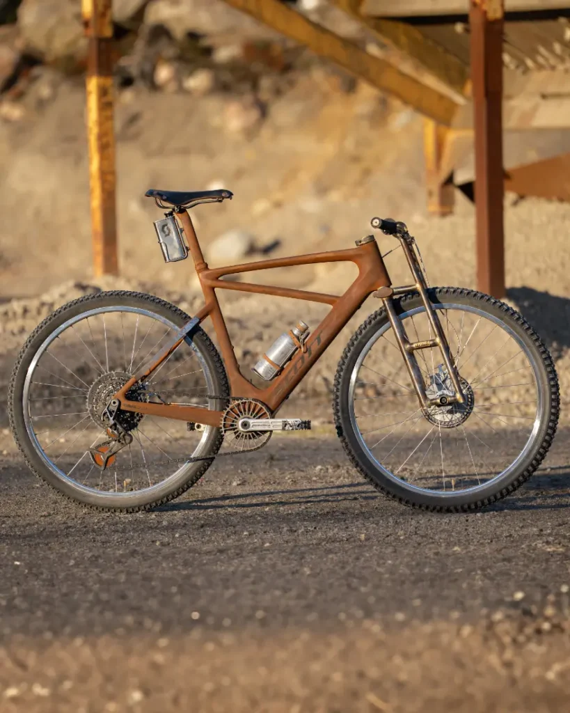 Una bicicleta de montaña vintage con un cuadro marrón, manillar plateado y neumáticos con tacos, que recuerda a un modelo SCOTT Solace, está estacionada sobre una superficie de grava cerca de una estructura de madera, evocando el encanto accidentado de un páramo indómito.