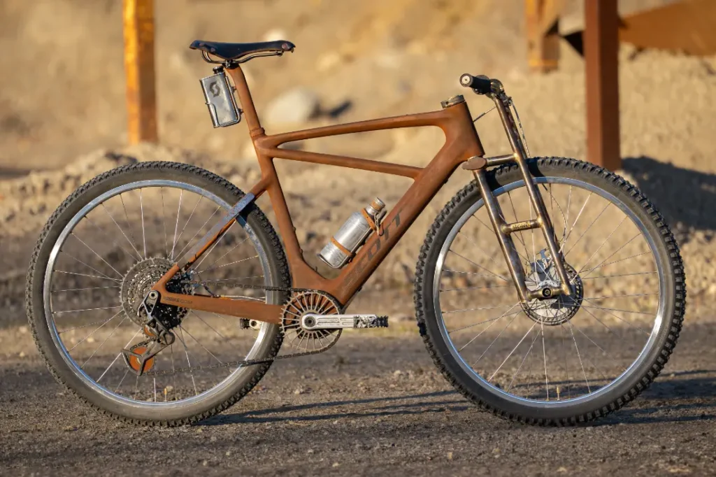 Una bicicleta de montaña SCOTT Solace, en un llamativo tono marrón, se encuentra confiadamente en un camino rocoso con dos botellas de agua adjuntas, lista para conquistar el páramo.