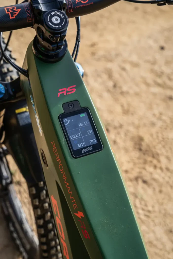 Primer plano de una bicicleta eléctrica verde con motor Polini E-P3+, con una pantalla digital que muestra la velocidad, la distancia, el porcentaje de batería y el modo de potencia sobre un fondo de arena. El diseño sofisticado muestra la potencia y precisión italianas en su máxima expresión.