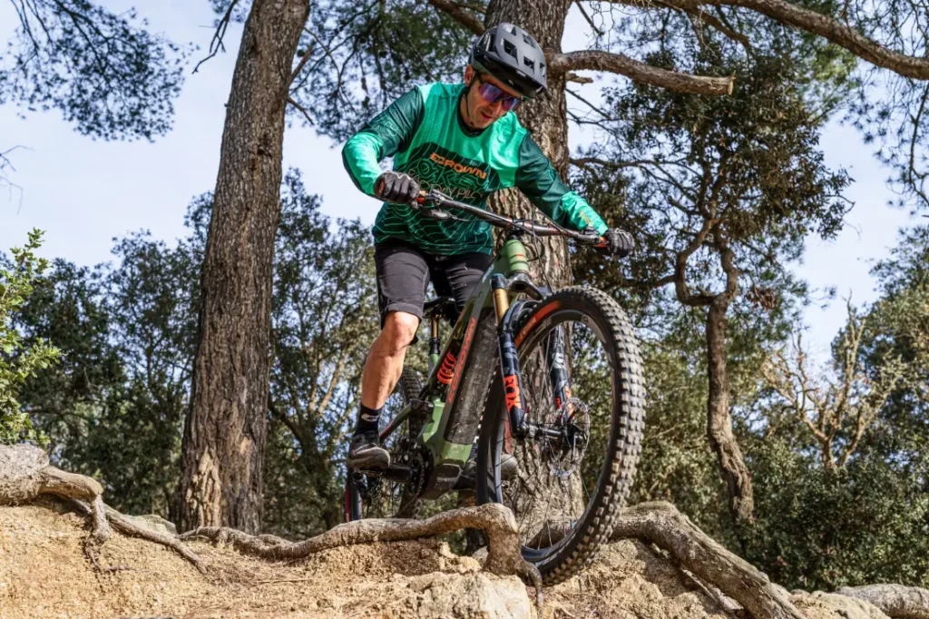 Una persona con un maillot verde y un casco recorre en bicicleta de montaña un terreno rocoso en un entorno forestal, impulsada por la impresionante Polini E-P3+ MX, que muestra pura potencia italiana.