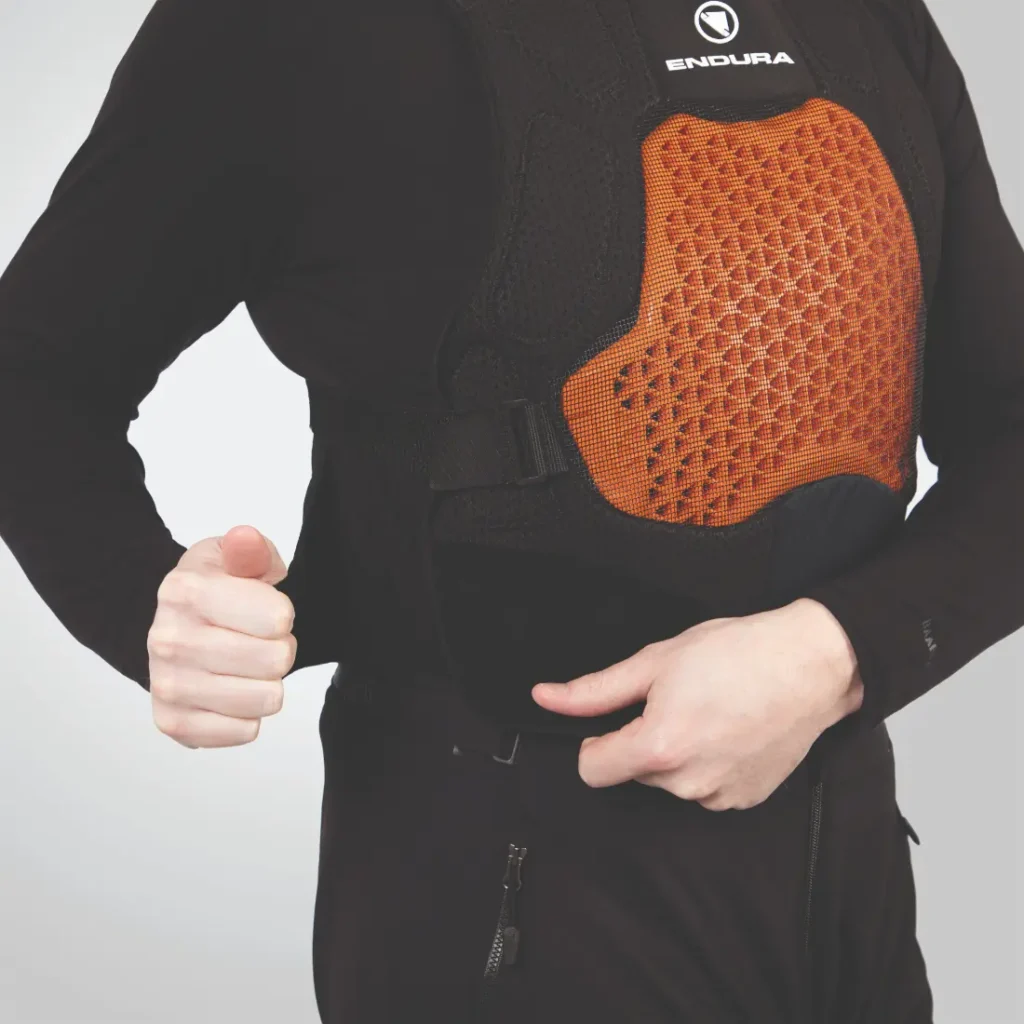 Una persona que viste un chaleco protector negro y naranja, ajustando la correa lateral con su mano derecha, muestra el diseño confiable de las protecciones Endura.