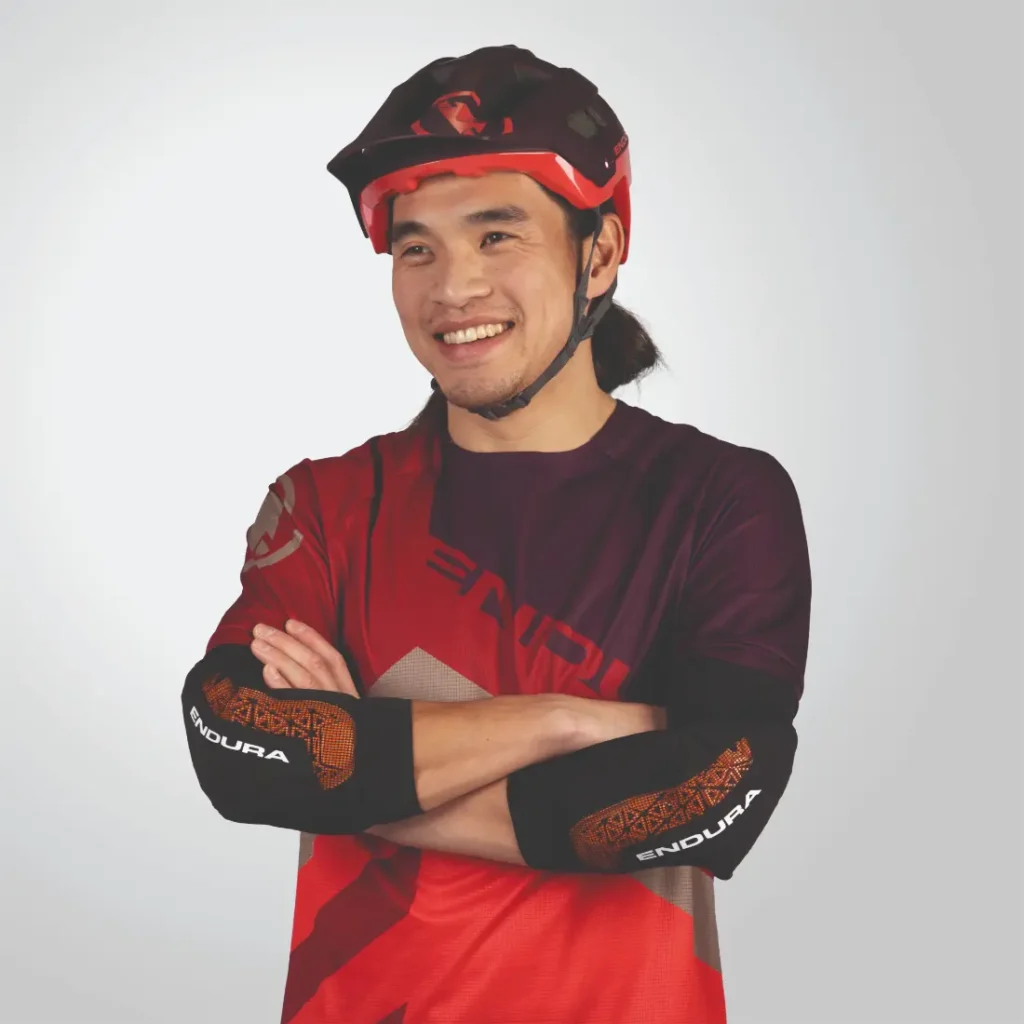 Una persona que lleva un casco rojo y una camiseta de ciclismo Endura, con los brazos cruzados y sonriendo sobre un fondo liso.