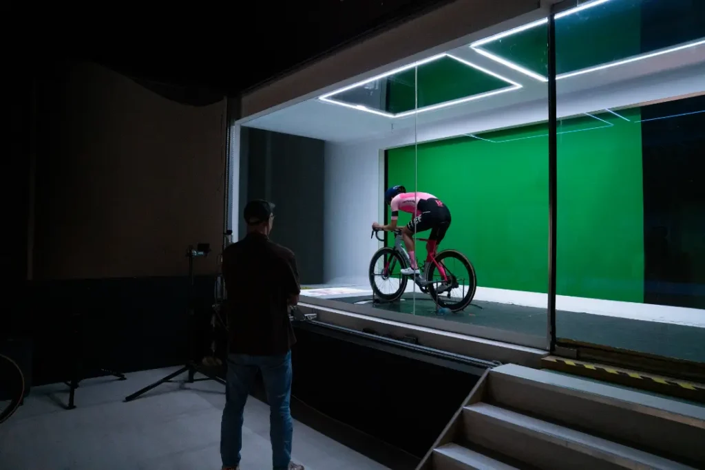 Un ciclista en una bicicleta fija, equipado con equipo POC Pro, pedalea enérgicamente en un estudio de pantalla verde, mientras una persona observa atentamente desde afuera del espacio, tal vez imaginando el futuro de las actuaciones de EF Pro Cycling.