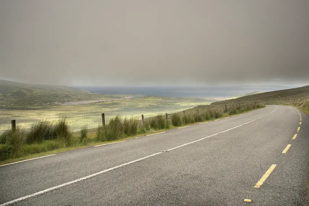 Un camino sinuoso con una línea discontinua amarilla serpentea a través del paisaje herboso de Conor Pass, enmarcado por un cielo nublado.
