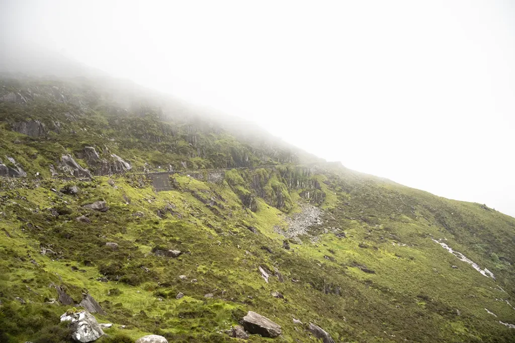 Una colina brumosa en Conor Pass, con exuberante césped verde y rocas dispersas, se encuentra bajo un cielo brillante y brumoso.