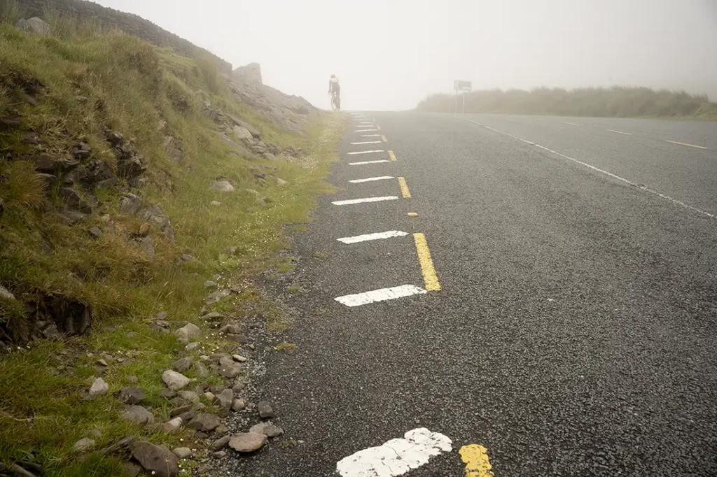 En una carretera con niebla cerca de Conor Pass, una persona anda en bicicleta a través de la niebla, flanqueada por un terreno cubierto de hierba y marcas viales visibles.