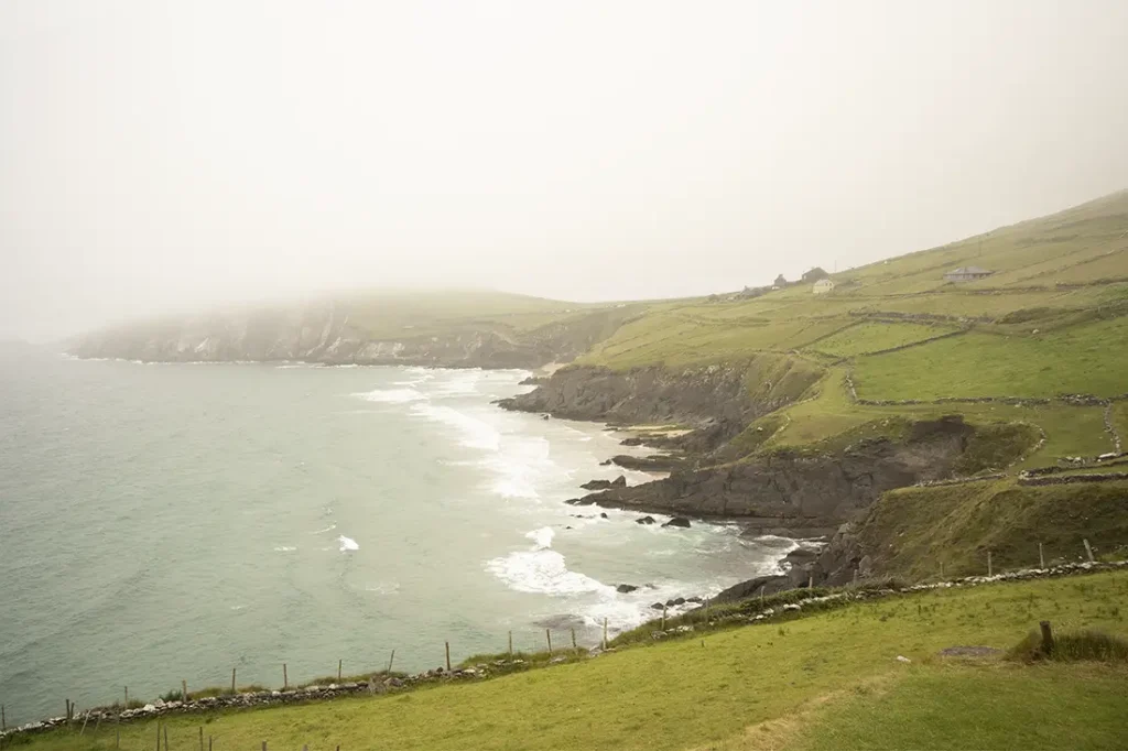 Paisaje costero brumoso con colinas verdes y acantilados que se unen al mar en Conor Pass. Las olas rompen suavemente contra la costa rocosa, mientras que a lo lejos se ven algunos edificios rurales.