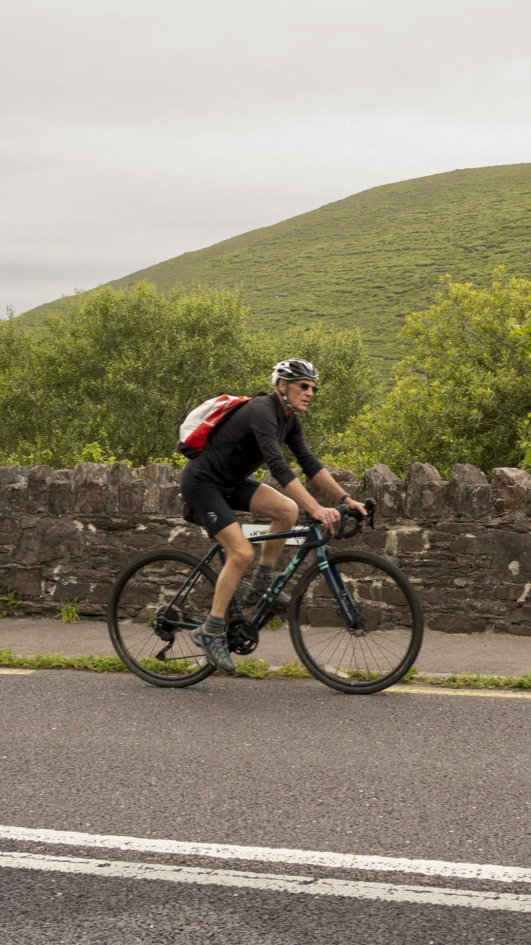 Un ciclista con casco y mochila recorre la ruta panorámica cerca de Conor Pass, pedaleando junto a un muro de piedra con una colina cubierta de hierba al fondo.