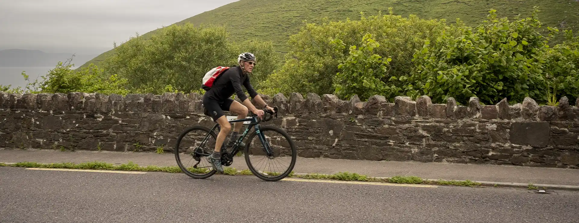 Un ciclista con una mochila roja recorre la pintoresca ruta de Conor Pass, recorriendo un camino bordeado por un encantador muro de piedra y una exuberante vegetación.