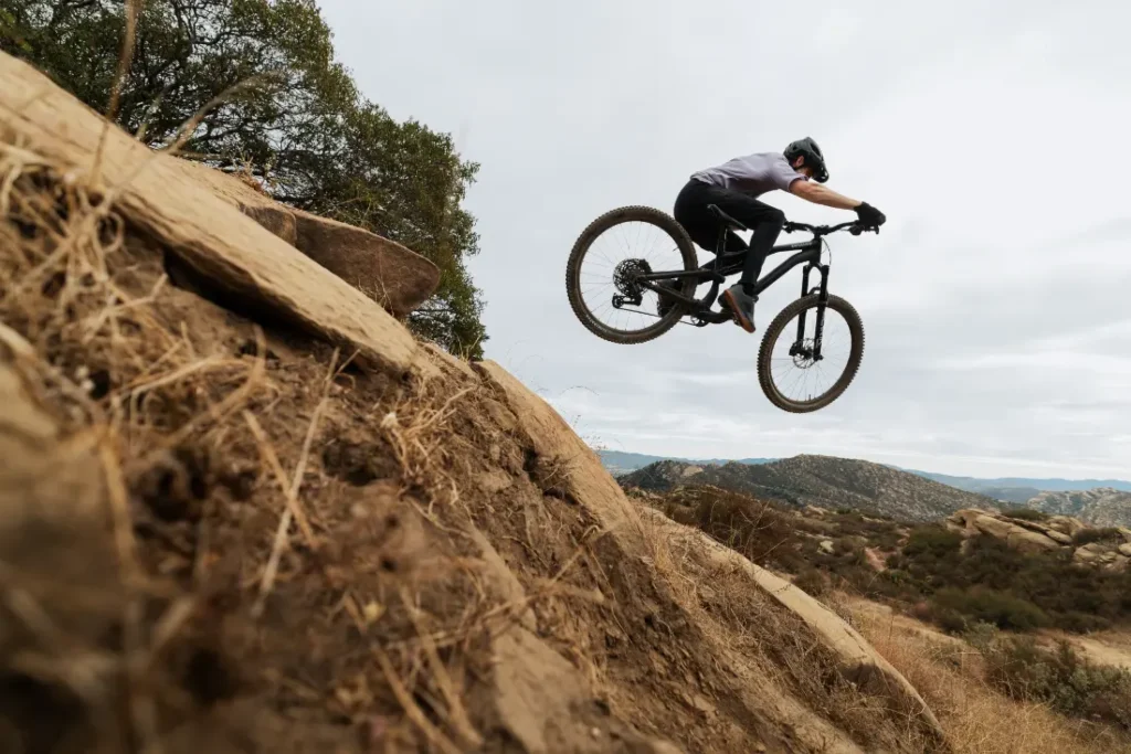 Un ciclista de montaña, impulsado por un SRAM Eagle 90, se eleva por los aires sobre un sendero rocoso, con colinas onduladas y un cielo nublado como telón de fondo.
