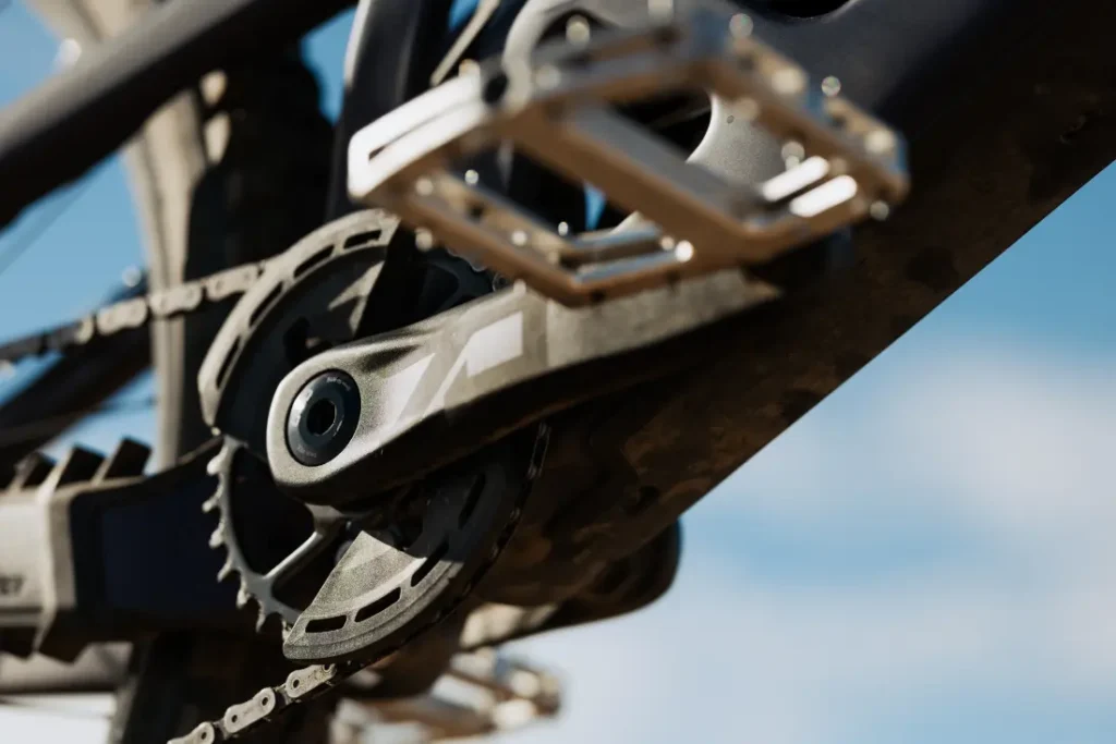 Primer plano de un juego de bielas y un pedal de bicicleta bajo un cielo azul claro, resaltando el plato SRAM Eagle 90 y los detalles de los cambios mecánicos.