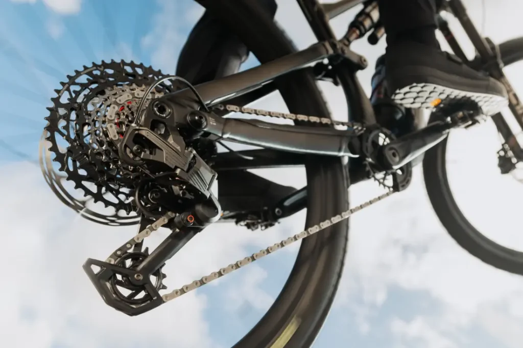 Primer plano del desviador trasero SRAM Eagle 70 y los cambios de una bicicleta mientras se conduce contra un cielo azul con nubes dispersas.