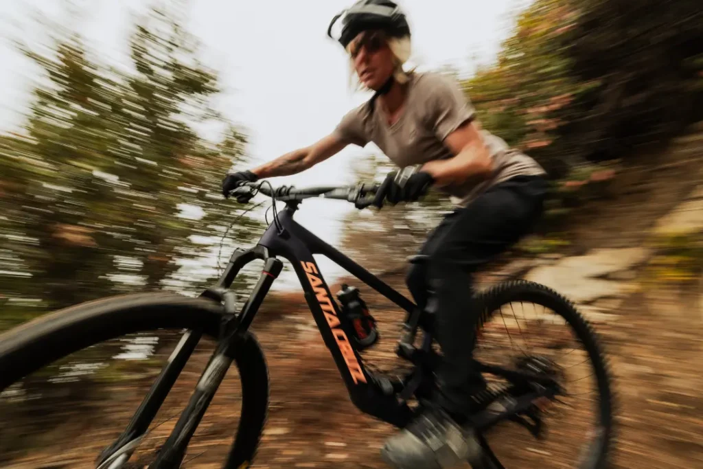 Una persona con casco y camisa gris monta una bicicleta de montaña con SRAM Eagle 90 en un sendero de tierra, rodeada de follaje borroso, lo que transmite una sensación de movimiento.