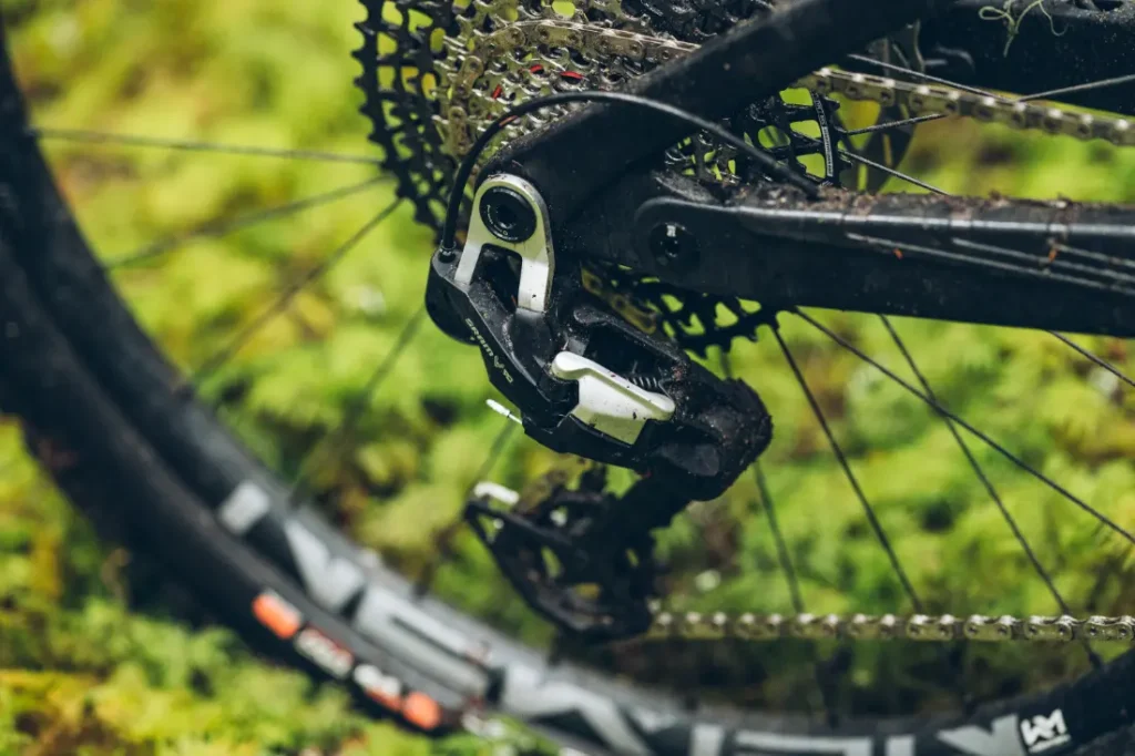 Primer plano del desviador trasero SRAM Eagle 90 y los cambios de una bicicleta sobre un fondo verde y herboso.