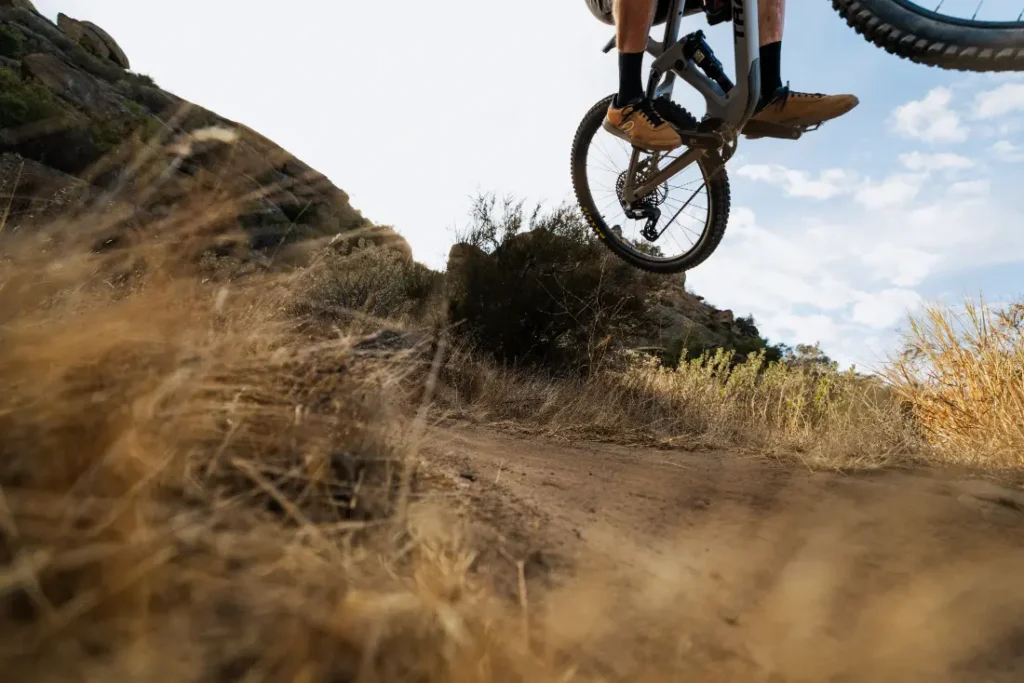 Un ciclista equipado con componentes SRAM Eagle 90 está en el aire, realizando un salto en un sendero de tierra. La hierba seca se extiende en primer plano, mientras que un cielo despejado se extiende al fondo, mostrando la emoción de la precisión y la aventura sobre dos ruedas.