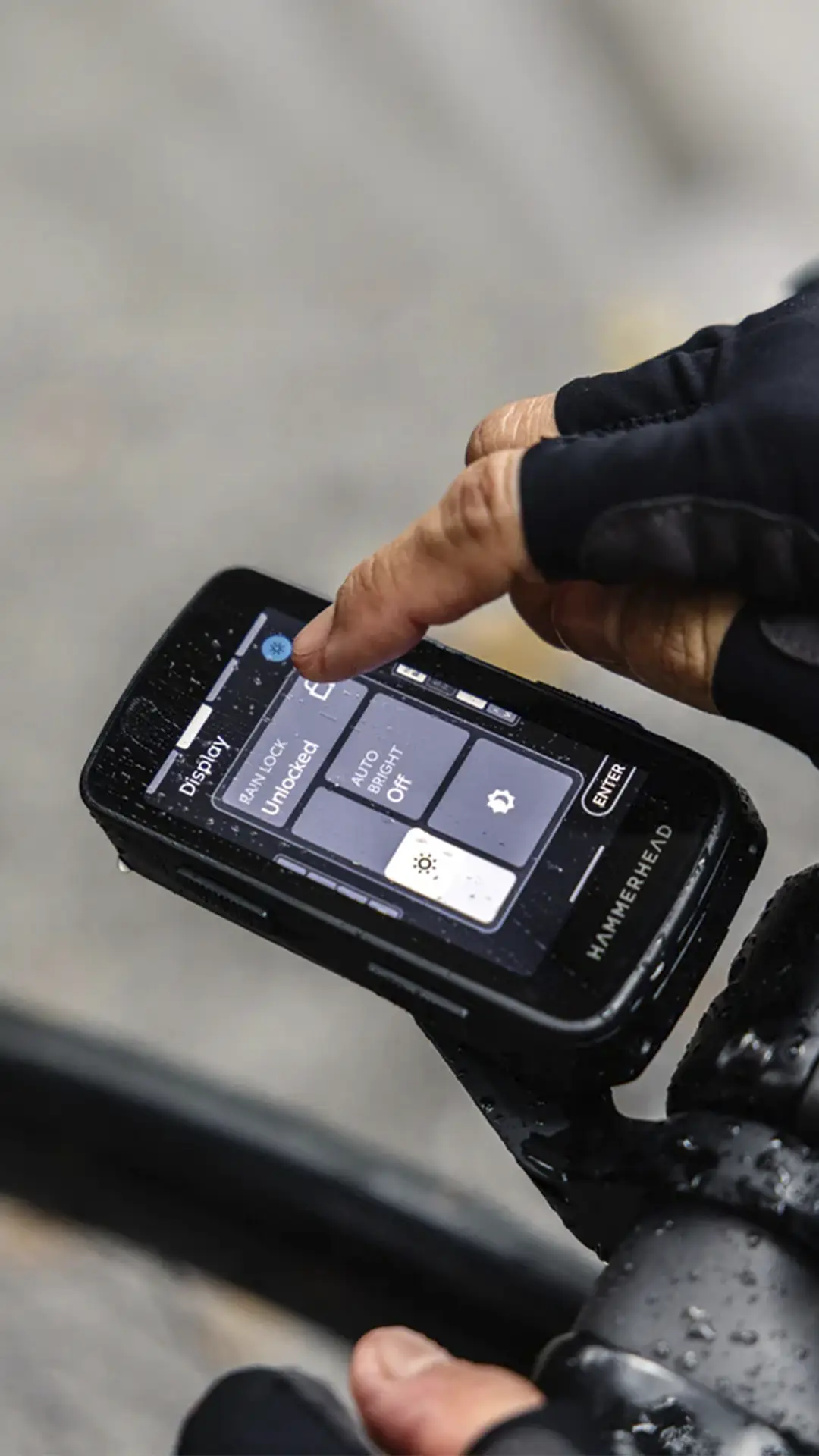 Una mano enguantada interactúa con un ciclocomputador con pantalla táctil montado en el manillar de una bicicleta, mostrando diversas configuraciones.