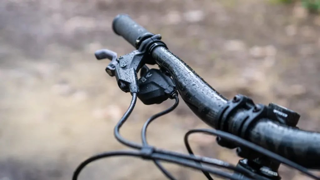 Primer plano de un manillar de bicicleta mojado, equipado con palancas de freno SRAM Eagle 90 y cables visibles sobre un fondo de camino exterior borroso.