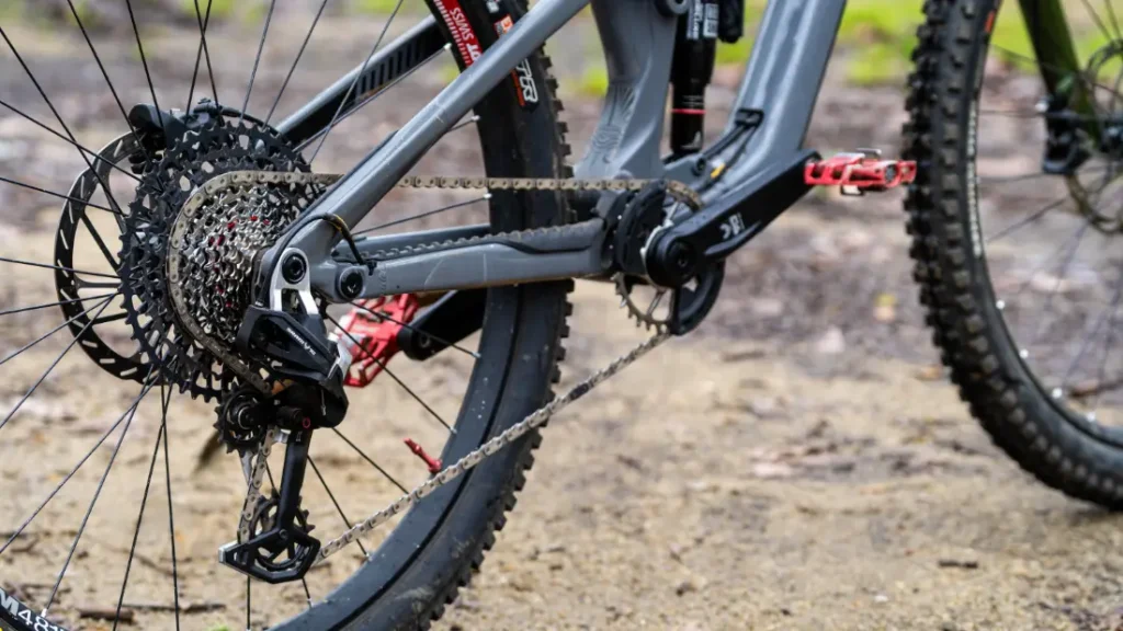 Primer plano de la rueda trasera de una bicicleta de montaña que muestra el cassette de cambios, el descarrilador y la cadena en un camino de tierra, con la precisión de los cambios mecánicos SRAM Eagle 70.