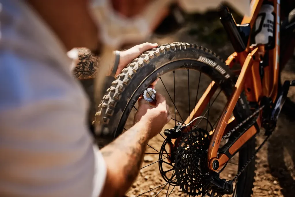 En un entorno exterior soleado, una persona utiliza un manómetro Unior para comprobar la presión de aire del neumático trasero de una bicicleta de montaña.