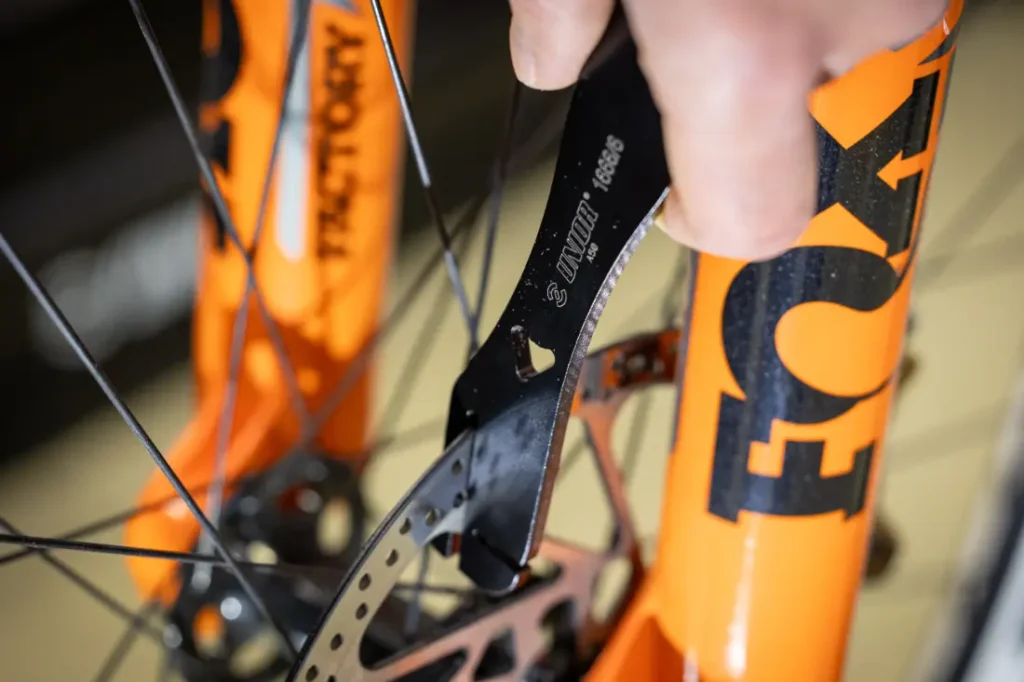 Una persona utiliza una herramienta Unior negra para ajustar el freno de disco en una horquilla de suspensión Fox naranja de una bicicleta.