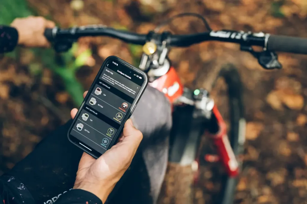 Una persona sostiene un smartphone con una aplicación de ciclismo mientras está sentada en una bicicleta de montaña roja en un bosque sereno. La MTB está equipada con funciones avanzadas como la RockShox Reverb AXS, que integra la tecnología de vanguardia Air-Over-Air para una experiencia de conducción mejorada.