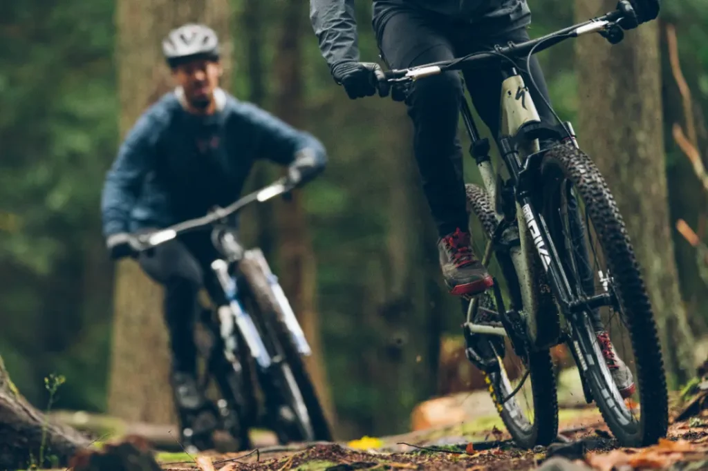 Dos ciclistas recorren una pista forestal en bicicleta de montaña, con sus ropas oscuras mimetizándose con el frondoso entorno. Equipados con la tecnología RockShox Reverb AXS, dominan cada curva con precisión, garantizando la seguridad con sus cómodos cascos.
