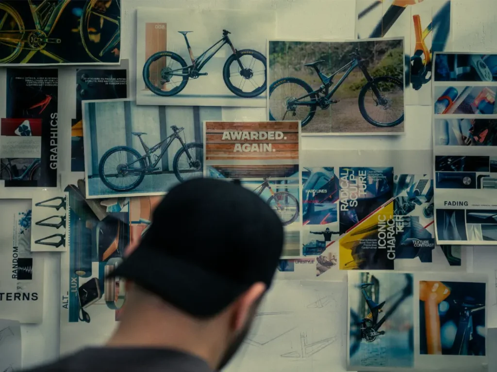 Una persona con una gorra negra se enfrenta a una pared adornada con imágenes de UNNO MITH, con fotografías de bicicletas y diversos materiales de diseño.