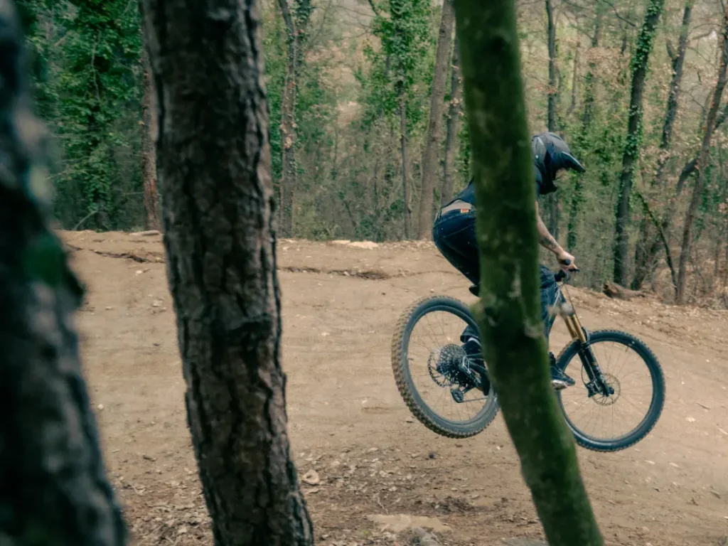 Ataviado con elegante equipo de ciclismo negro, un ciclista que luce la UNNO MITH se sube con destreza a una bicicleta de montaña por un sendero de tierra. El exuberante bosque de fondo, con sus imponentes árboles y su vibrante follaje, realza la emoción del paseo.