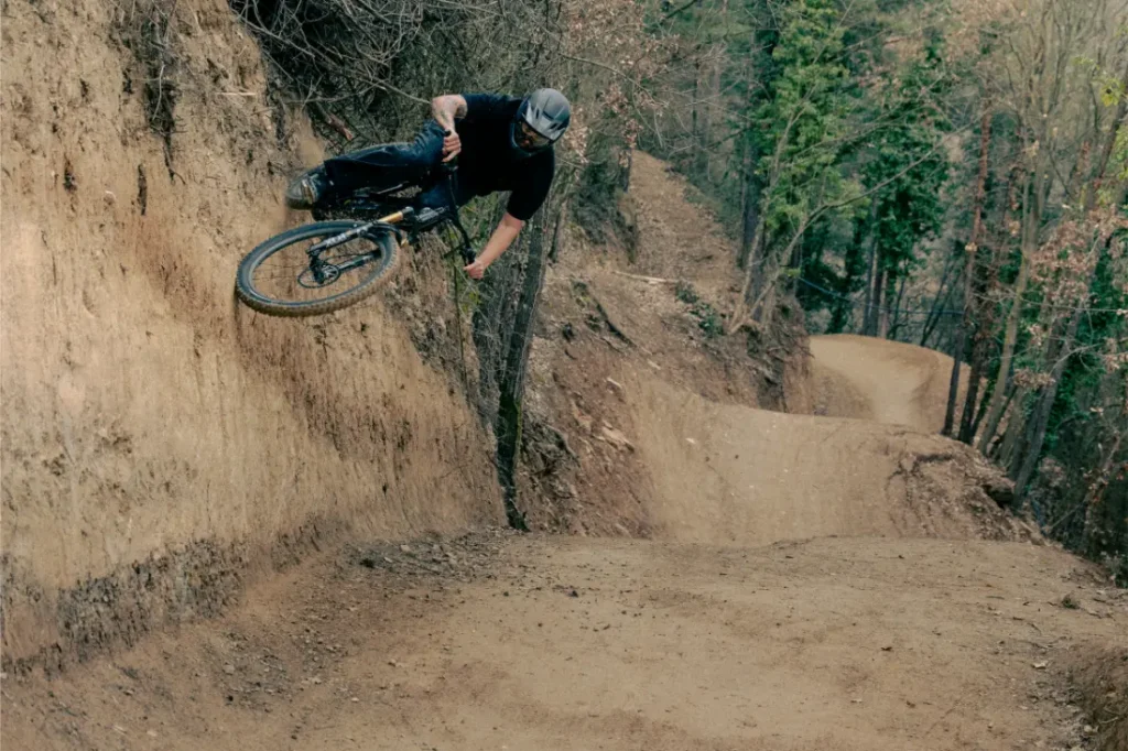 Un ciclista de montaña en un UNNO MITH realiza con gracia un recorrido por la pared en un sendero de tierra, rodeado de árboles imponentes.