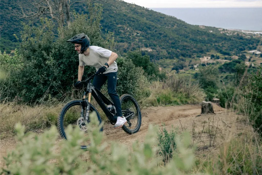 Un ciclista con casco maniobra con destreza una bicicleta de montaña UNNO MITH a lo largo de un sinuoso sendero de tierra, rodeado de exuberantes colinas verdes con el mar sereno extendiéndose más allá.