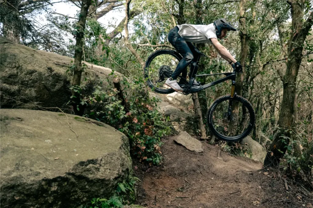 Un ciclista de montaña en una bicicleta UNNO MITH realiza un atrevido salto desde una roca en una zona boscosa, demostrando habilidad y precisión mientras viste casco y equipo de protección.
