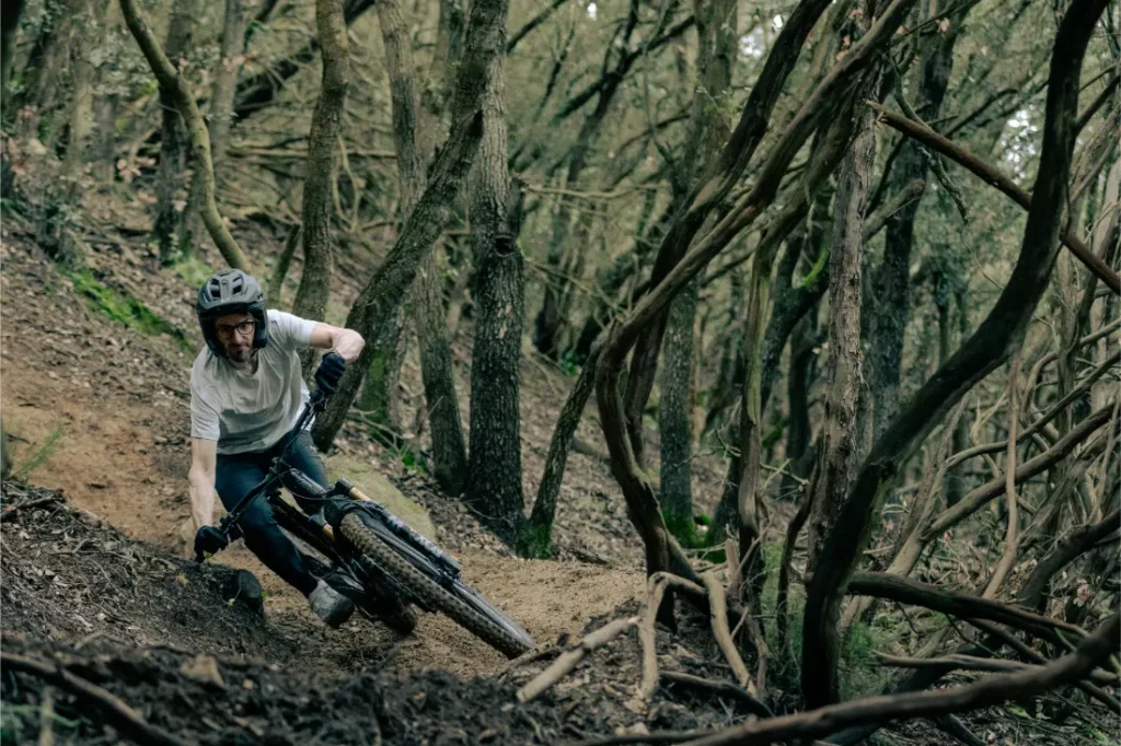 Un ciclista con camisa blanca y casco recorre con destreza un sendero de tierra estrecho y empinado a través de un bosque denso, apoyándose fuertemente en una bicicleta de montaña UNNO MITH.
