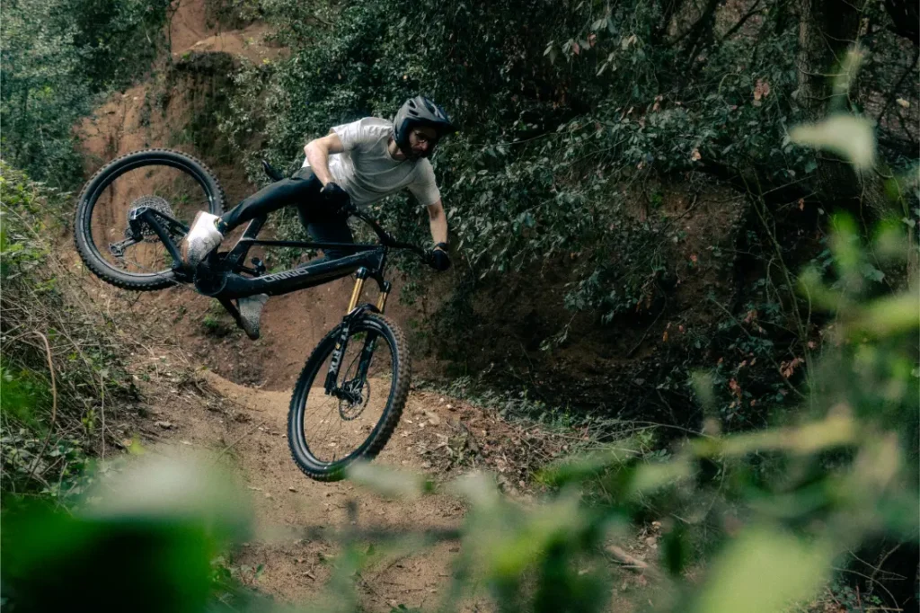 Un ciclista de montaña en un UNNO MITH se eleva por los aires, ejecutando un salto en un sendero de tierra, envuelto de exuberantes árboles y follaje.
