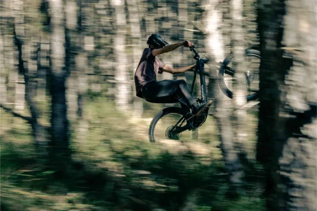 Una persona realiza con confianza un caballito en su bicicleta de montaña UNNO MITH, maniobrando con gracia a través del bosque mientras los árboles se difuminan en un fondo lleno de movimiento.