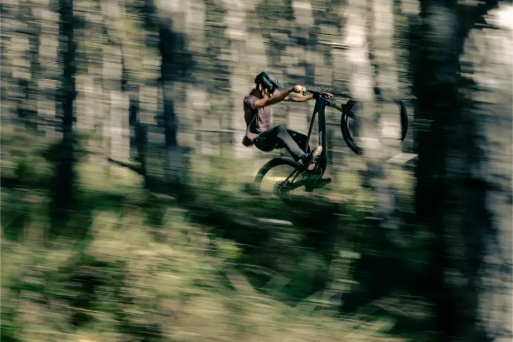 Una persona que lleva un casco realiza un salto en una bicicleta de montaña UNNO MITH en un bosque, con el desenfoque de movimiento capturando la velocidad.