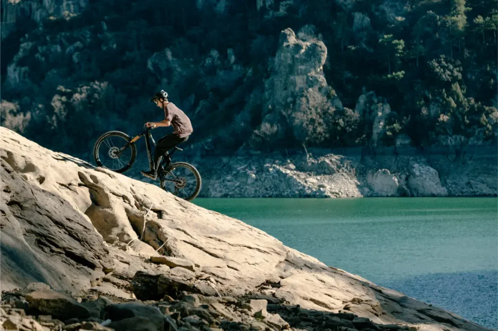 Una persona que lleva casco monta sin esfuerzo en una bicicleta de montaña UNNO MITH por una pendiente rocosa junto a un lago tranquilo, con un acantilado boscoso que crea un telón de fondo impresionante.