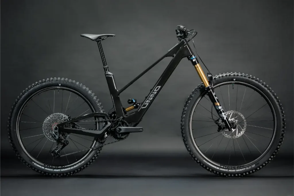 La UNNO MITH, una elegante bicicleta de montaña negra con un cuadro de suspensión total, frenos de disco y neumáticos con tacos, está ingeniosamente ubicada sobre un fondo gris oscuro.