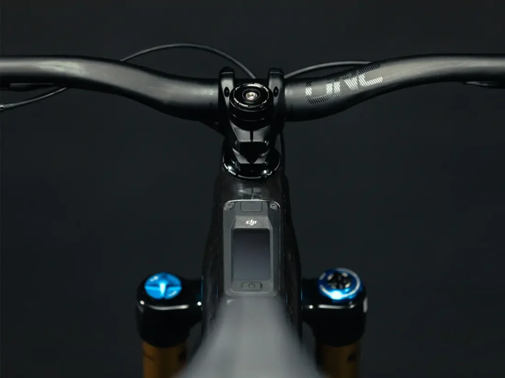 Primer plano de un manillar y una potencia de bicicleta UNNO MITH con empuñaduras negras y una pequeña pantalla en el centro, sobre un fondo oscuro.