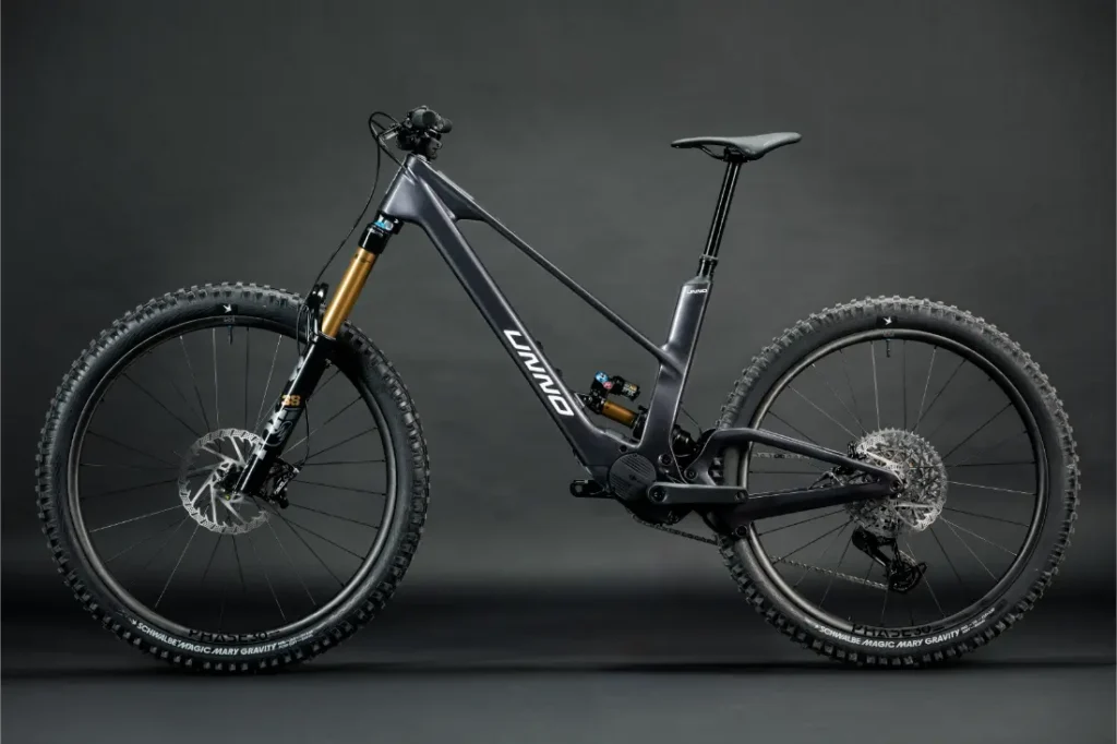 La bicicleta de montaña negra UNNO MITH cuenta con horquillas de suspensión grandes, neumáticos anchos y un cuadro elegante, resaltado sobre un fondo oscuro y liso.