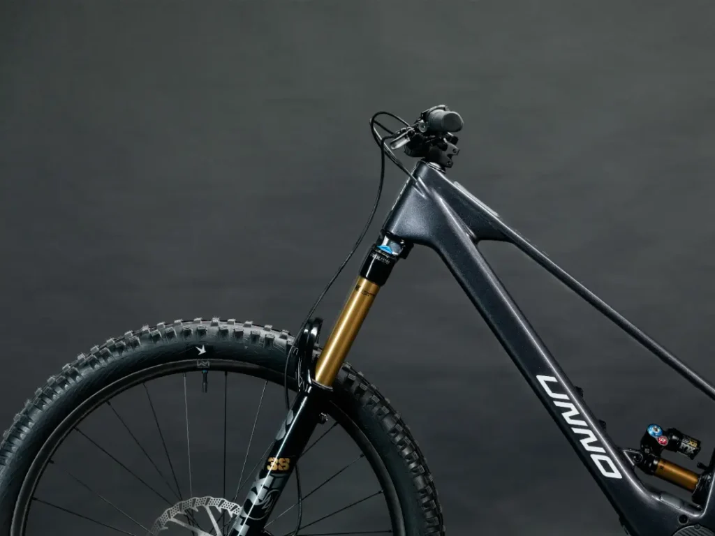 Vista de cerca de una bicicleta de montaña UNNO MITH negra con suspensión delantera, neumáticos con tacos y manillar, sobre un fondo oscuro.