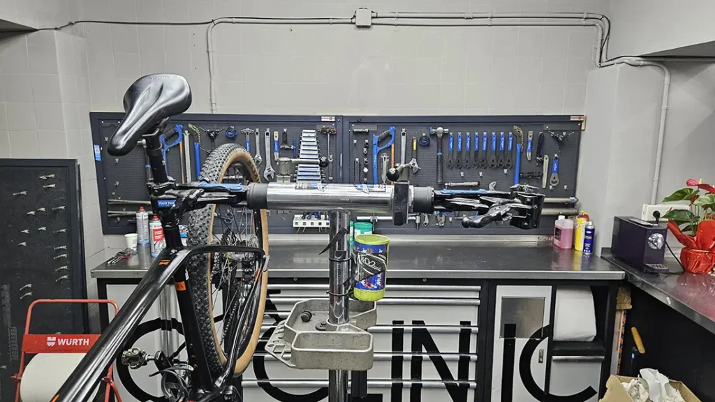 Una bicicleta está montada en un soporte de reparación en el taller de Biciclinic, con varias herramientas organizadas en la pared detrás y suministros de mantenimiento en el mostrador.
