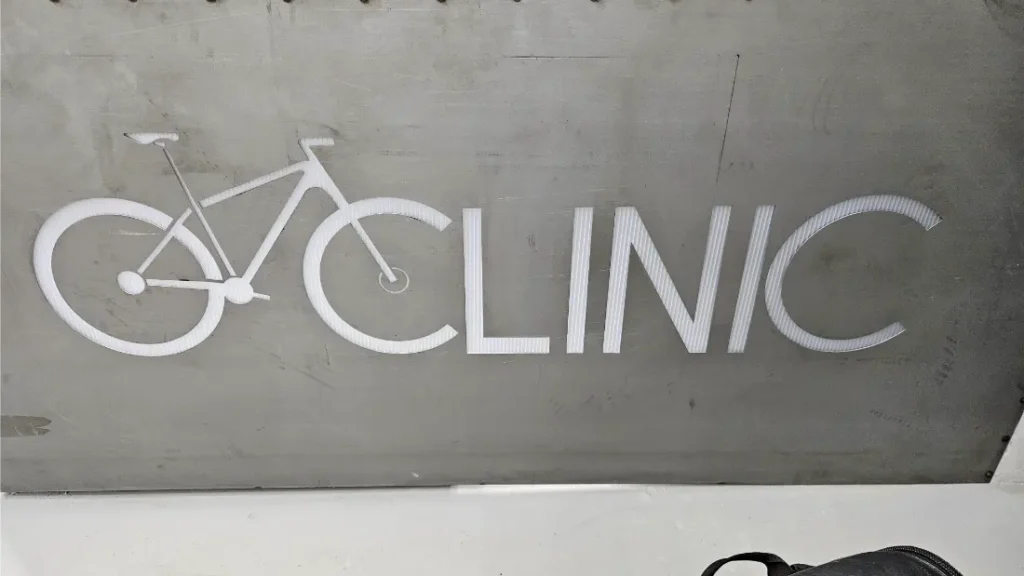 Un cartel con la palabra "Biciclinic", donde la letra "C" está estilizada como el diseño de una rueda y un cuadro de bicicleta, se destaca sobre un fondo gris.